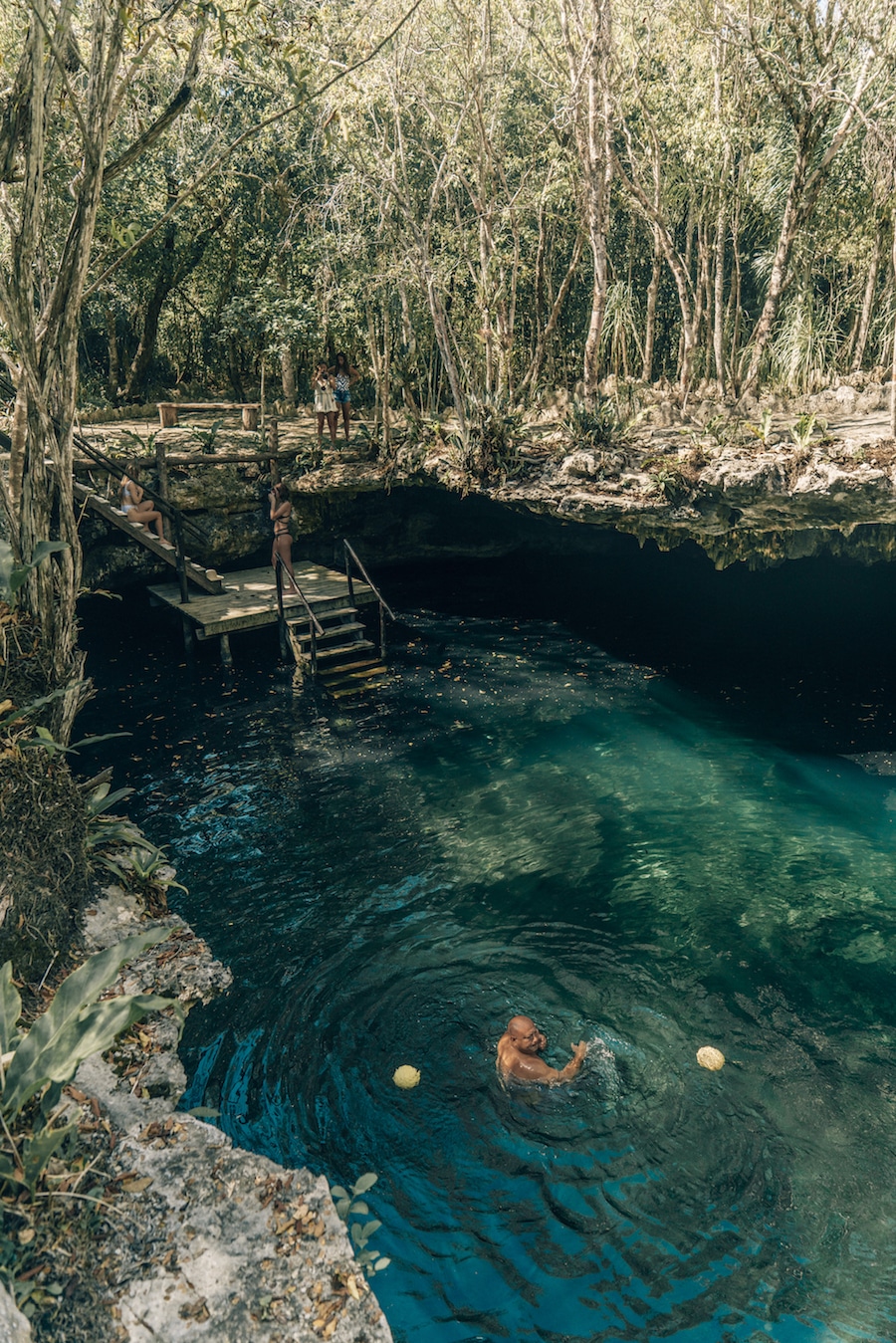 A man swimming in the middle of the clear blue waters of a cenote while few people are on the wooden staircase surrounded by lush vegetation 