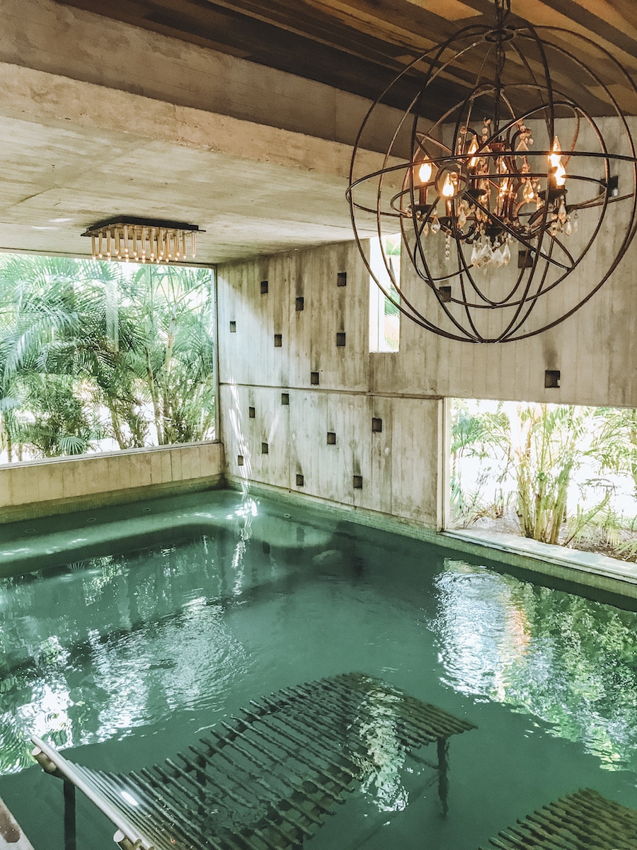 A clear indoor pool with two submerged sun loungers and a rustic chandelier with large windows and clear views of the outdoors
