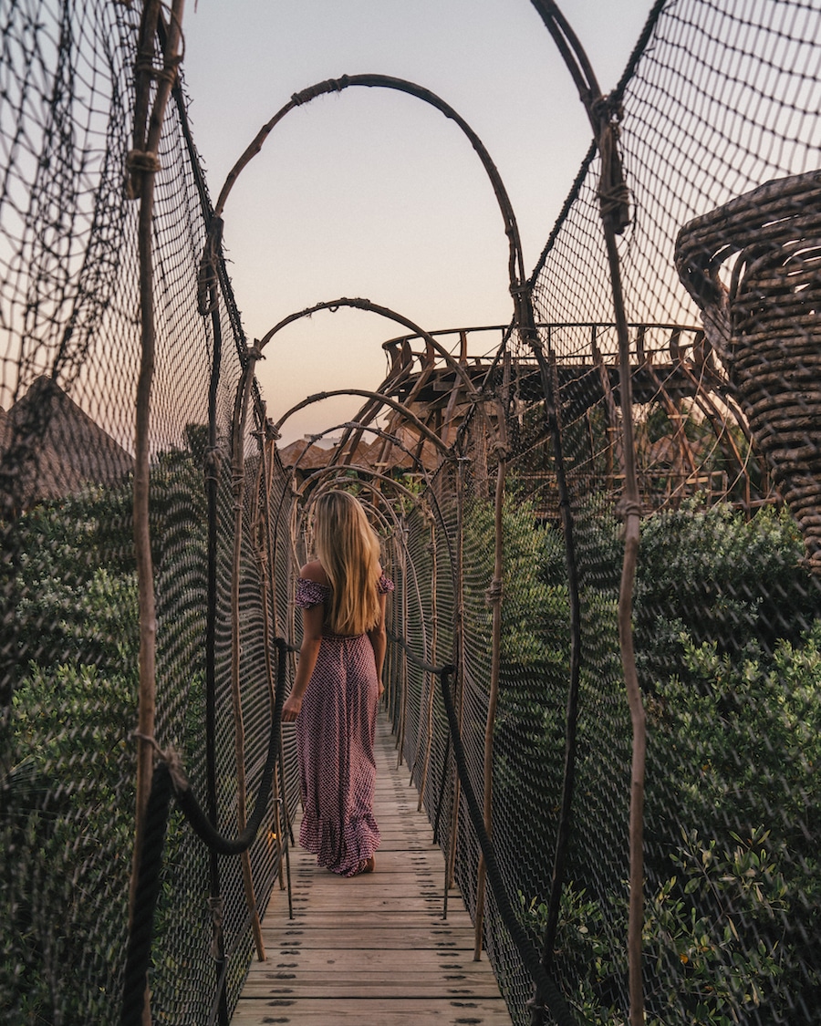 Me in a dress in the middle of a narrow wooden bridge surrounded by green trees under sunset skies