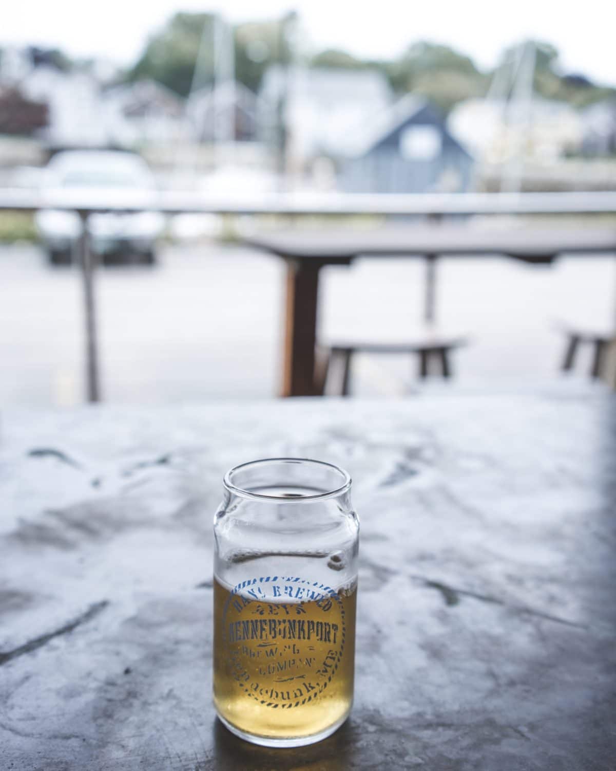 A glass of beer with fading print on a table with a blurry view of the outdoors through a glass window.