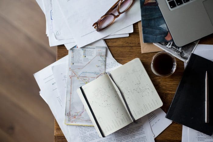 A wood desk with a journal, maps and coffee spread out on top.