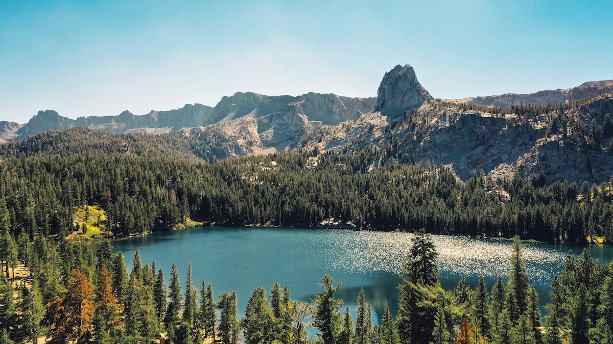Jagged mountains in the distance with a stunning blue lake in front of it surrounded by pine trees.