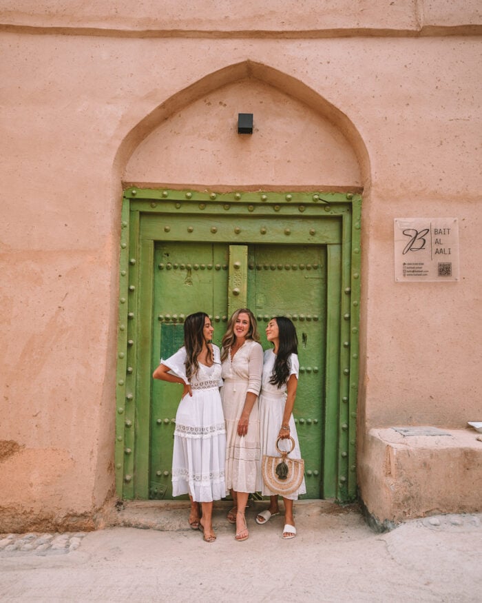 Catarina Mello, Michelle Halpern and Shelbi Okumura in Al Hamra Mud Village in front of green doorway on their Oman road trip itinerary 