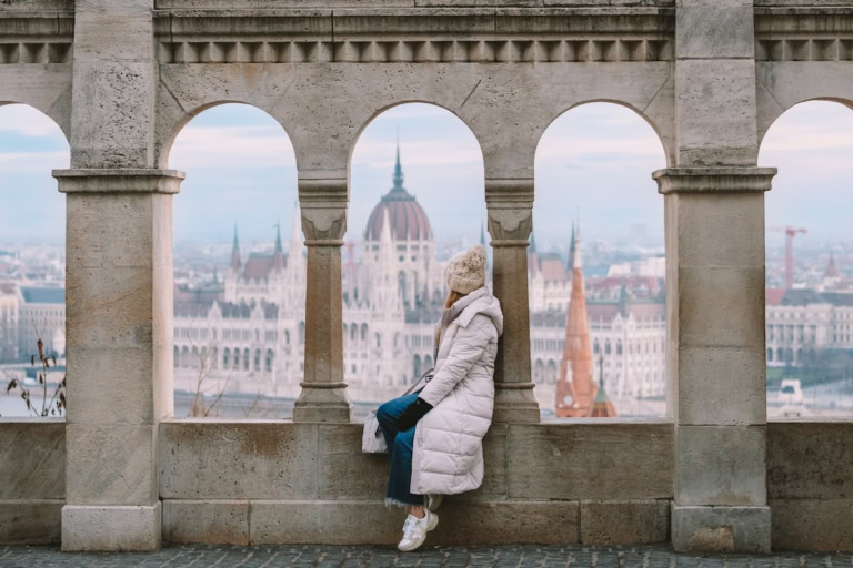Michelle Halpern sitting at Fisherman's Bastion in Budapest with a big white coat on looking out at the view.