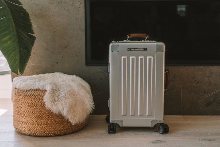Aluminum 40L Sterling Pacific carry-on bag sitting next to a sheepskin covered pouf and in front of a fireplace
