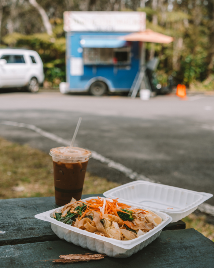 noodle dish A white container with a noodle dish and a coffee sitting on a picnic table. It features a blue food truck in the background.