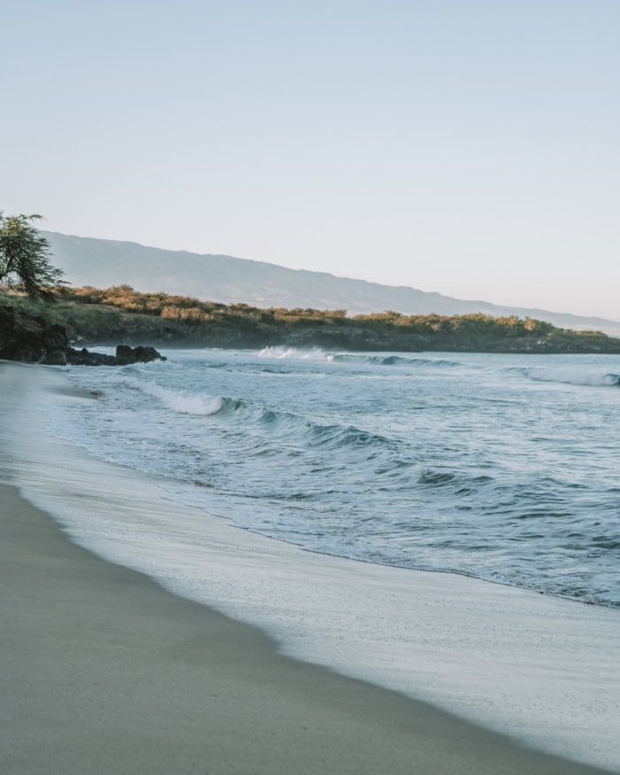 Hapuna Beach Beach at sunrise in front of the Westin Hapuna Beach resort. The sand in the image is gray and the water is a beautiful light blue color.