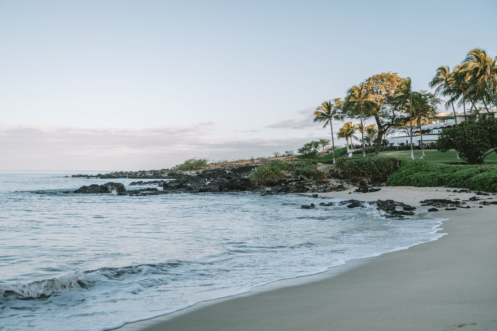 Westin Hapuna Beach Beach at sunrise in front of the Westin Hapuna Beach resort. The sand in the image is gray and the water is a beautiful light blue color.
