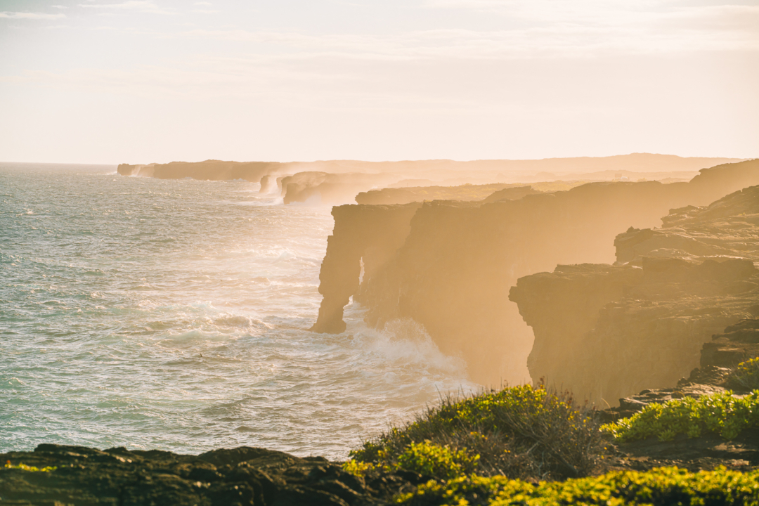Dramatic ocean cliffs at a sunset on the Big Island of Hawaii. The image features waves crashing below and golden light illuminating the shore.
