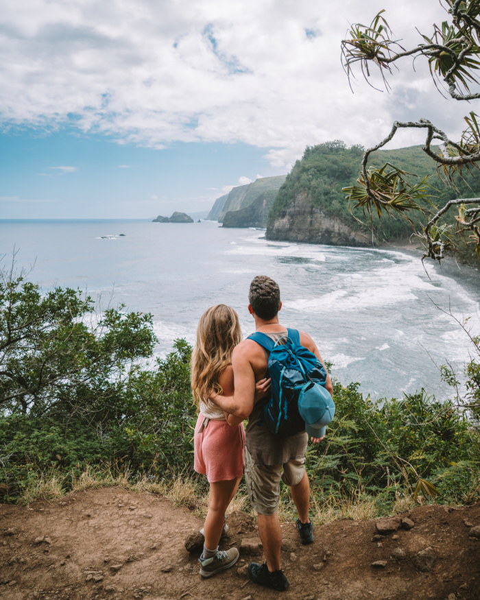 JT and Michelle trail JT and Michelle overlooking the ocean standing on a trail in Big Island, Hawaii. They are surrounded by plants and cliffs in the background.
