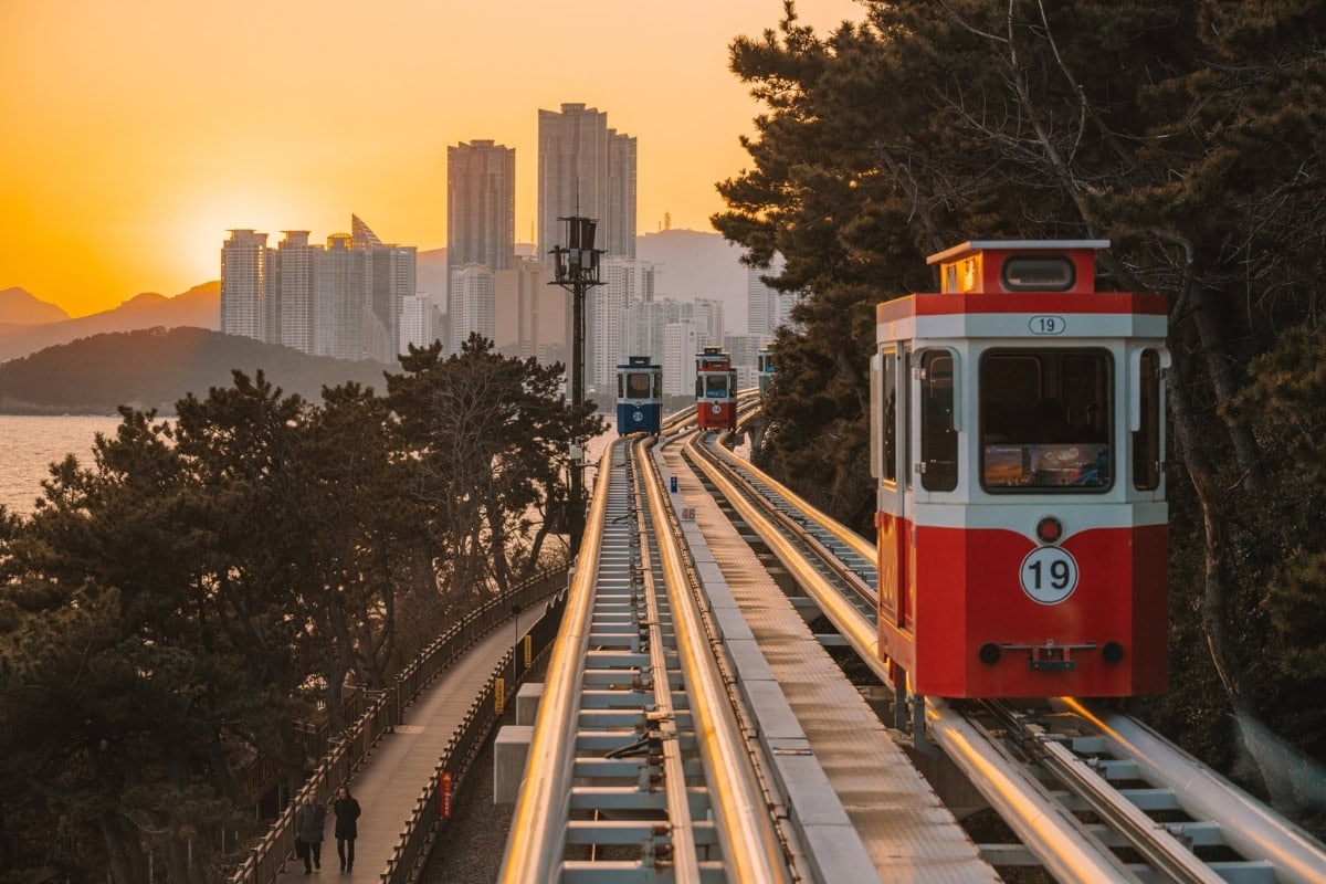 Red train car on elevated tracks at sunset with city buildings in the background.