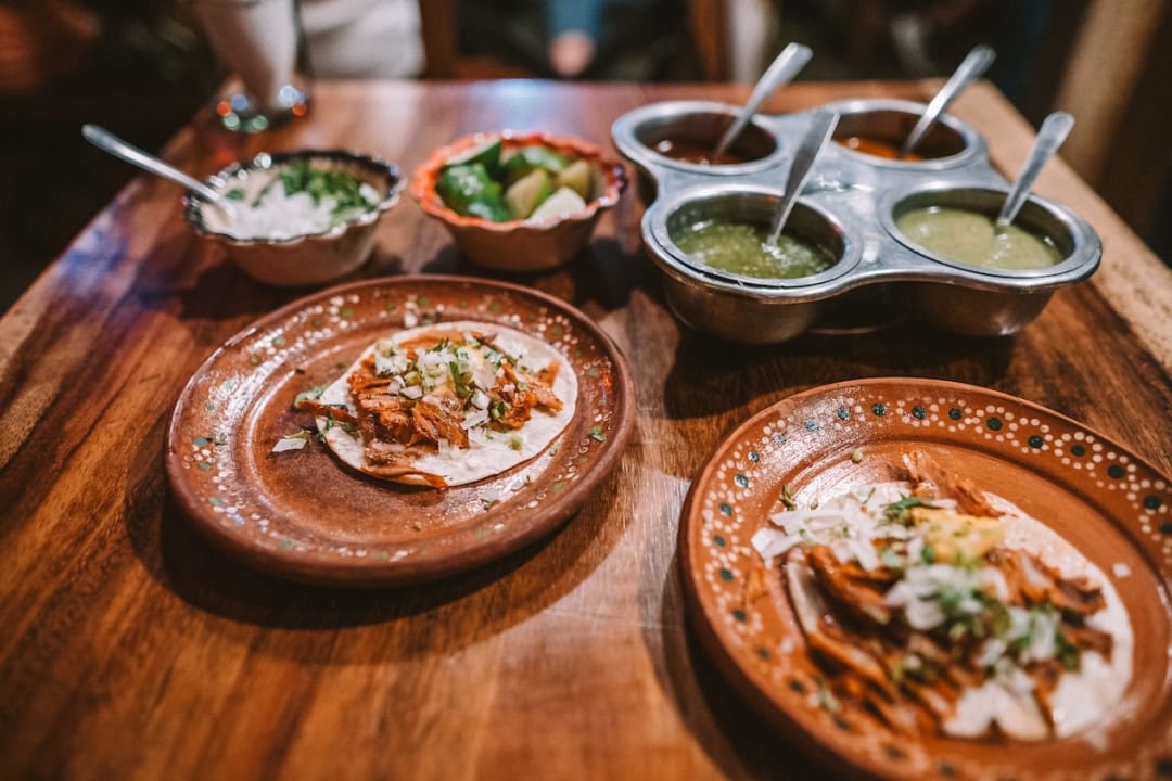 Two plates of tacos on a wooden table with a bunch of condiments and salsas in a carrier behind it.