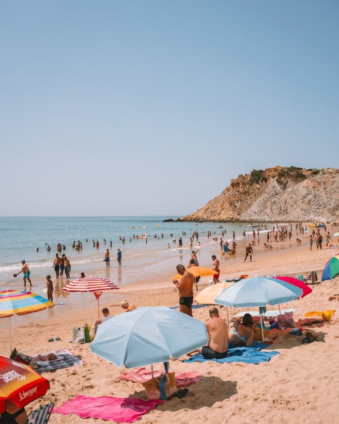 Algarve Portugal Itinerary Colorful umbrellas line the beach in summer as tons of beachgoers enter shallow water.
