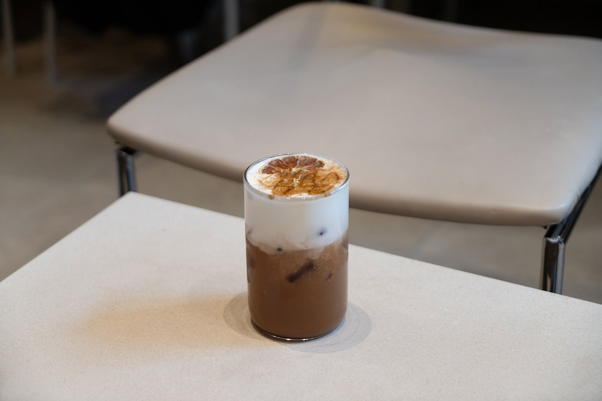 coffee with orange on top Iced beverage sitting on a table that has foam at the top and a dried slice of orange
