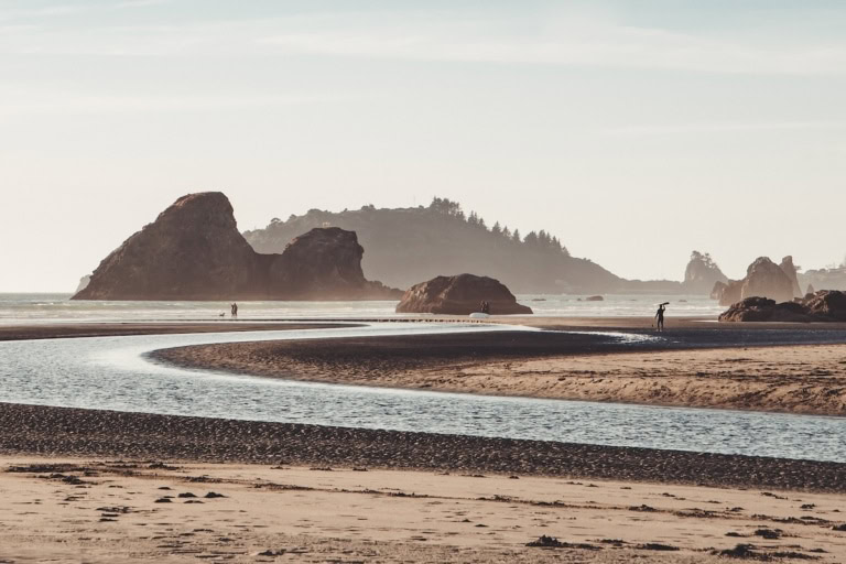 Golden hour at Moonstone Beach in Trinidad - there's a stream running through the beach and a surfer walking out.