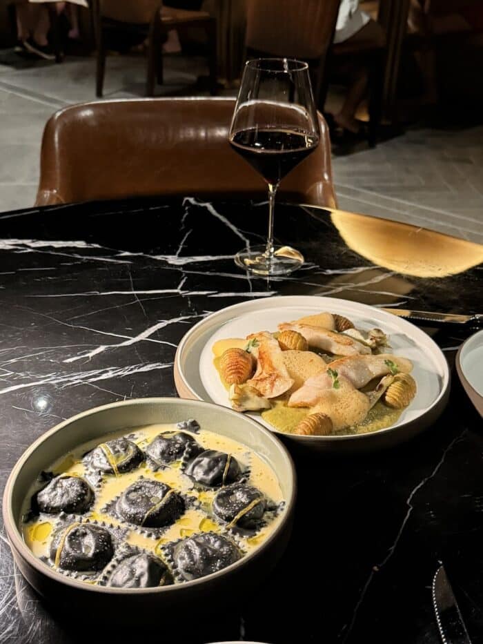 Two pasta dishes on a black marble table with a glass of red wine
